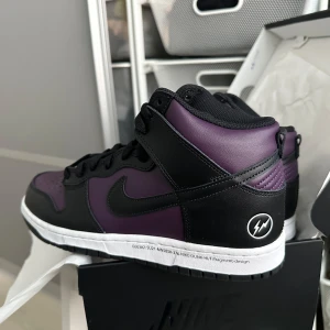 Nike Dunk High Fragment Design svart/lila 42.5 - Nike Dunk High i samarbete med Fragment Design. Skorna har svart och lila ovandel i skinn, vit mellansula och svart yttersula. Fragment-logga på hälen och klassisk Nike Swoosh på sidan. Perfekta för dig som gillar unika sneakers med streetwear-känsla.