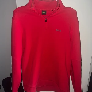 Hugo boss tröja - Säljer en röd långärmad tröja från BOSS med halv dragkedja framtill och diskret logga på bröstet. Tröjan har en sportig look och en randig detalj på ena ärmen. Perfekt för dig som gillar stilrena plagg med en twist.