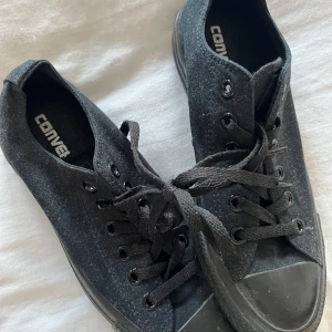 Svarta Converse sneakers - Klassiska svarta Converse sneakers med lågt skaft och svarta snören. Skorna har en platt sula och är tillverkade i textil med gummidetaljer på tån och sulan. Perfekta för en avslappnad och stilren look.