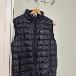Svart dunväst från EA - Säljer en svart quiltad dunväst från EA med hög krage och dragkedja framtill. Västen har en stilren design med EA-logga på bröstet och passar perfekt för lager-på-lager-stil.