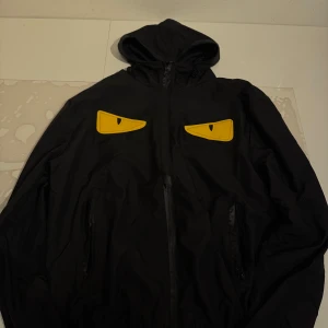 Svart vindjacka med gula ögon-detaljer - Svart vindjacka med huva och dragkedja framtill. Två gula ögon-detaljer på bröstet ger jackan en unik look. Fickor med dragkedja på sidorna. Perfekt för dig som vill sticka ut.
