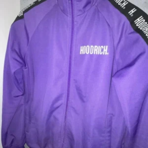 Lila track jacket från Hoodrich - Säljer en lila track jacket från Hoodrich med dragkedja och logga på bröstet. Jackan har svarta band med vit text längs axlarna och hög krage. Perfekt för en sportig och streetwear-inspirerad stil.