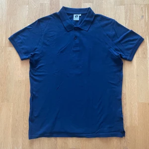 Mörkblå pikétröja från HangTen - Säljer en klassisk mörkblå pikétröja från HangTen med krage och två knappar framtill. Tröjan är kortärmad och tillverkad i mjuk bomull, perfekt för en stilren och avslappnad look samt matchar väldigt bra till shorts eller jeans! 