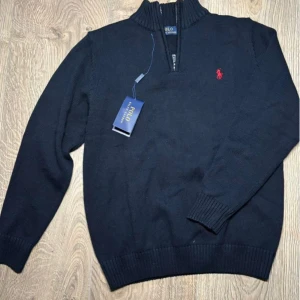 Ralph Lauren half zip blå - Mörkblå stickad tröja från Polo Ralph Lauren med halv dragkedja och röd broderad logga på bröstet. Tröjan har lång ärm och ribbade muddar vid ärmslut och nederkant. Perfekt för lager-på-lager-stil.
