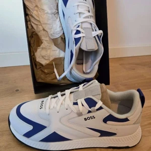 Vita och blå sneakers från BOSS - Snygga sneakers från BOSS i vitt och blått med sportig design. Skorna har snörning, rund tå och platt sula. Perfekta för en trendig och avslappnad stil.