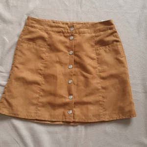 Brun mockakjol med knappar från H&M Divided - Säljer en brun A-linjeformad mockakjol från H&M Divided. Kjolen har silvriga knappar framtill och två fickor. Perfekt att styla med en enkel topp eller stickad tröja.