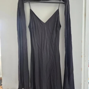 Svart långklänning med snörning från Yue London - Elegant svart långklänning från Yue London med smala axelband och djup v-ringning. Klänningen har öppen rygg med dekorativ snörning och pärldetaljer. Tillverkad i polyester och acetat, följsam och glansig känsla. Perfekt för speciella tillfällen.