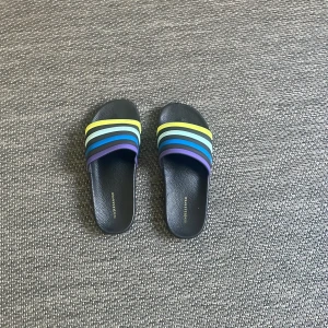 Svarta sandaler med färgglada ränder från Reserved - Säljer ett par svarta sandaler från Reserved med breda remmar i blått, grönt, gult och lila. Perfekta för sommaren och riktigt sköna att glida runt i. Snygg design med platt sula och öppen tå.