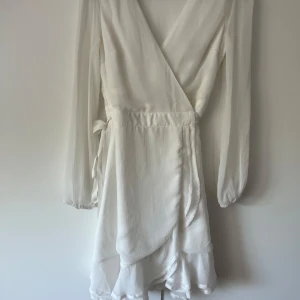 Student klänning - Säljer en vit omlottklänning från Bubbleroom med volangdetaljer nertill, använd 1 gång men ser ut som ny. Klänningen har v-ringning och knytband i midjan för en smickrande passform. Perfekt för sommarens tillfällen och framförallt till skolavslutning eller student. Midjeviddenär ungefär 65-70cm. Bystvidden är 82cm!