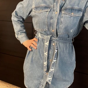 Jeansklänning med bälte från Calvin Klein - Snygg jeansklänning från Calvin Klein med knappar framtill och två bröstfickor. Klänningen har ett avtagbart bälte med CK-logga som markerar midjan. Perfekt för en avslappnad men trendig look.