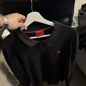 Svart pikétröja med dragkedja från Hugo - Säljer en svart pikétröja från Hugo med klassisk krage och diskret dragkedja framtill. Tröjan har lång ärm och liten röd logotyp på bröstet. Perfekt för en stilren och enkel look.