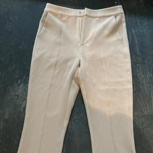 Beige kostymbyxor med slits - Snygga beige kostymbyxor med raka ben och diskreta slitsar nertill framtill. Byxorna har hög midja och pressveck längs benen för en stilren look. Perfekta att matcha med en skjorta eller stickad tröja! Byxorna är köpta i London och är i storlek S