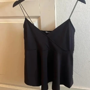 Svart omlott-linnetopp med tunna axelband - Säljer en svart omlott-linnetopp med tunna axelband och v-ringning. Toppen har en lätt utsvängd passform och är perfekt för varma dagar. Enkel och stilren design som passar till mycket.