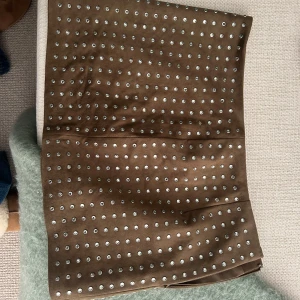 Brun mocka kjol med nitar - Säljer en ny aldrig använd brun kjol med silvriga nitar över hela tyget. Kjolen har en rak passform och är tillverkad i ett mockaliknande material. Större storlek så den passade mig perfekt lowrise. Super gullig för sommarfest! 