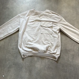 Beige sweatshirt med bröstficka från MON - Säljer en beige sweatshirt från MON i storlek S. Tröjan har en rund halsringning, långa ärmar och en praktisk bröstficka med lock. Mjuk bomullskänsla och ribbade muddar vid ärmslut och nederkant.