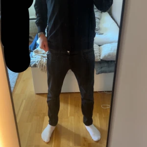 Svarta jeansbyxor från Replay - Säljer ett par svarta jeansbyxor från Replay med klassisk femficksdesign och dragkedja. Byxorna har en slim passform och är tillverkade i ett hyper stretchigt material som sitter snyggt på benen. Modellen på bilden är 185 och jeansen är lite korta