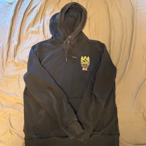 Svart hoodie med tryck från Morris - Svart hoodie från Morris med huva och dragsko. Tryck med Morris-logga och brittisk flagga på bröstet. Känguruficka framtill och mjukt material, perfekt för en avslappnad stil.