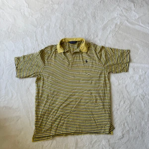 Gul och marinblå randig pikétröja från Polo Golf Ralph Lauren - Säljer en klassisk gul pikétröja med marinblå ränder från Polo Golf Ralph Lauren. Tröjan har korta ärmar, krage och knäppning vid halsen. Tillverkad i 100% bomull och passar perfekt för en varm golfrunda 😊