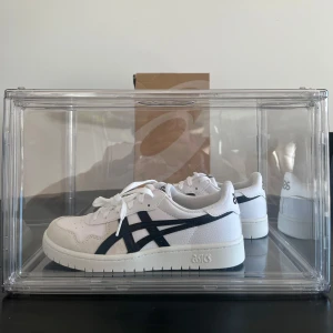 Asics sneakers med svarta detaljer - Säljer ett par fräscha vita Asics sneakers med svarta ränder på sidorna. Skorna har klassisk låg profil, rund tå och platt sula. Perfekta för en clean och sportig stil. Materialet är syntet och textil.