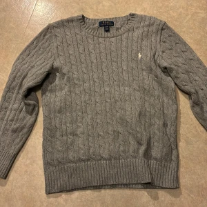 Grå stickad tröja från Polo Ralph Lauren - Säljer en klassisk grå stickad tröja från Polo Ralph Lauren med kabelstickat mönster och rund halsringning. Tröjan har lång ärm och den ikoniska broderade loggan på bröstet. Perfekt för lager-på-lager-stil.