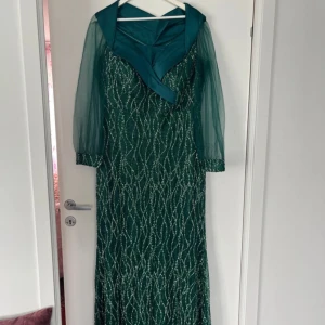 Grön långklänning med glittrigt mönster och sjalkrage - Elegant grön långklänning med glittrigt mönster och transparenta ärmar. Klänningen har en bred sjalkrage i satinliknande material och ett bälte i midjan. Perfekt för speciella tillfällen. Kommer med justerbar kjol.