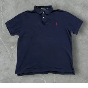 Mörkblå pikétröja från Ralph Lauren - Klassisk mörkblå pikétröja från Ralph Lauren med korta ärmar och krage. Tröjan har två knappar framtill och den ikoniska röda loggan broderad på bröstet. Perfekt för en stilren och avslappnad look.
