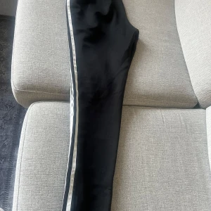 Svarta Adidas mjukisbyxor med vita ränder - Svarta mjukisbyxor från Adidas med klassiska vita ränder längs sidorna och Adidas-logga på höften. Byxorna har en sportig look och är perfekta för en avslappnad stil.