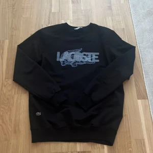 Svart sweatshirt från Lacoste - Svart sweatshirt från Lacoste med klassisk krokodil-logga och text framtill i grått. Tröjan har rund halsringning och långa ärmar. Perfekt för en avslappnad och stilren look. Liten i storlek xl men passar för Medium