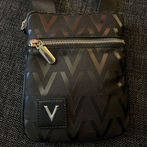 Svart axelväska från Mario Valentino - Snygg svart axelväska med mönster av blanka V och silverfärgade dragkedjor. Väskan har justerbar axelrem och två fack med dragkedja. Märkeslogga framtill och diskret präglad etikett inuti. 