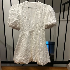 Vit kortklänning med puffärm och broderade blommor - Superfin vit kortklänning med puffiga ärmar och v-ringning. Klänningen har broderade blommor över hela tyget och en lätt ballongformad nederkant. Perfekt för sommaren eller student! ❤️