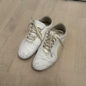 Vita sneakers från Puma med snörning - Säljer ett par vita sneakers från Puma med klassisk logga på sidan och hälen. Skorna har snörning och rund tå, samt detaljer i syntetmaterial. Perfekta för en sportig och stilren look.