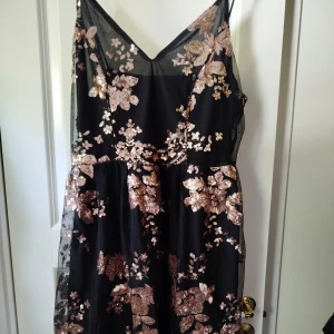 Svart klänning med paljettblommor från SHEIN - Svart klänning från SHEIN med tunna axelband och v-ringning. Klänningen är dekorerad med stora blommor i roséguldiga paljetter och har dragkedja i sidan. Perfekt för festliga tillfällen. Storlek XL Shein curve. Endast använd ett fåtal gånger.