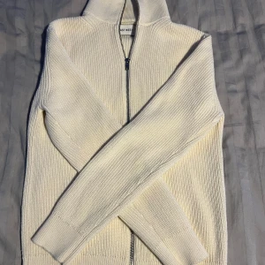 Vit stickad kofta med dragkedja från East West - Säljer en stilren vit stickad kofta från East West. Tröjan har hel dragkedja framtill, hög krage och ribbad struktur. Perfekt för lager-på-lager och enkel att matcha med olika outfits. Koftan är näst intill ny andvänd att fåtal gånger.