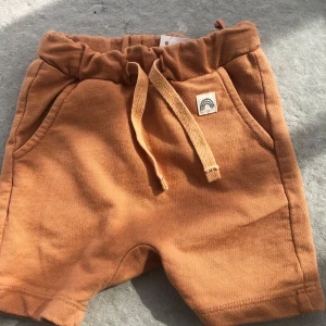 Orange shorts med snörning och regnbågsmärke - Säljer ett par orange shorts med resår och snörning i midjan. Shortsen har två fickor och en liten patch med regnbågsmotiv framtill. Perfekta för varma dagar.