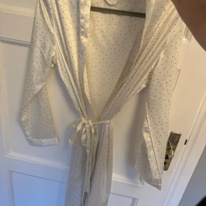 Vit prickig ”silkesmorgonrock” - Prickig morgonrock i silkesliknande material. Denna är aldrig använd så inga slitningar på något sätt. Den stängs med ett band men har också ett ”innerband” för att hålla ännu bättre. Ej äkta silke. Normal i storleken men passar även S.