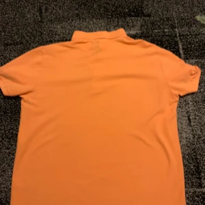 Orange pikétröja från Lacoste - Säljer en orange pikétröja från Lacoste med klassisk krage och korta ärmar. Tröjan har den ikoniska gröna krokodilloggan broderad på bröstet och knappar vid halsen. Den är i helt ny har aldrig använt den. Perfekt för en stilren och sportig look. Pris kan diskuteras!