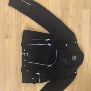 Svart jacka från ZARA med bälte - Säljer en svart skinnjacka från ZARA med silvriga dragkedjor och bälte i midjan. Jackan har flera fickor med dragkedja och klassisk krage. Perfekt för en cool och stilren look.