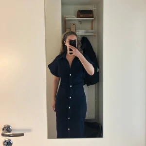 Marinblå klänning med knappar - Snygg marinblå klänning med knappar längs hela framsidan. Klänningen har korta ärmar och en figurnära passform som framhäver siluetten. Perfekt för dig som gillar stilrena och klassiska plagg. Prislappen är också kvar, den är endast provad.