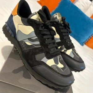 Valentino Rockrunners - Säljer ett par Valentino Rockrunner sneakers med svart, grått och beige camouflage-mönster. Skorna har svarta snören, detaljer i mocka och skinn samt en orange häl. Perfekta för dig som vill sticka ut med stil.