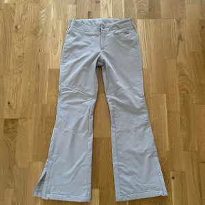 Beige friluftsbyxor från Columbia - Beige/gråa byxor från Columbia med broderad logga vid benslutet. Byxorna har bootcut-modell, dragkedja i benslutet och en ficka framtill. Perfekta för utomhusaktiviteter och har en bekväm passform.                            Innerbenslängd: 57cm, lårvidd: 23cm, fotvidd: 26cm
