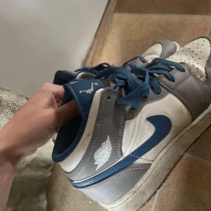 Nike Air Jordan 1 Low i blått och grått - Säljer ett par Nike Air Jordan 1 Low sneakers i blått, grått och vitt. Skorna har klassisk snörning, rund tå och platt sula. Perfekta för dig som gillar streetwear och vill ha en ikonisk sneaker i din samling.