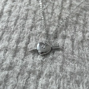 Auric Amour - Silver Heart & Arrow Pendant - Silverpläterat halsband i stainless steel med hjärta och pil, dekorerat med glittrande stenar. Justerbar längd 44 + 4 cm. En tidlös symbol för kärlek med modern elegans