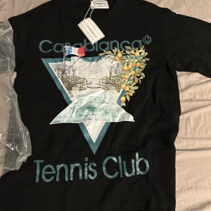 Svart Casablanca Tennis Club t-shirt - Svart t-shirt från Casablanca med stort grafiskt tryck framtill som visar en tennisbana, franska flaggan och apelsinträd. Texten 'Tennis Club' i blått. Rund hals och korta ärmar. Tillverkad i 100% bomull, perfekt för dig som gillar sportig och unik stil. Passar även storlek M alltså Sizeup.