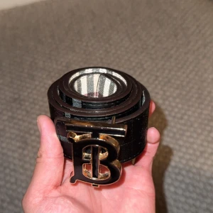 Burberry bälte med rutig design - Snyggt Burberry bälte med klassiskt rutig mönster i beige, svart, vitt och rött. Bältet har en bred form och en stor metallspänne med Burberrys ikoniska TB-logga i guld och svart. Tillverkat i skinn och perfekt för att lyfta vilken outfit som helst. ( 2st sidor/2st färger)