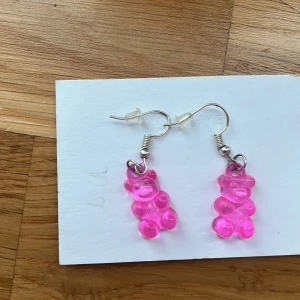 Rosa gelébjörnar örhängen - Supergulliga örhängen med rosa gelébjörn-berlock i plast. Perfekt för dig som vill ha något lekfullt och färgglatt! Hängande modell med silvriga krokar och transparenta pluppar. Sött och trendigt accessoar som ger din look en rolig twist. Jag själv är nickelallergiker och har inte haft några problem. 