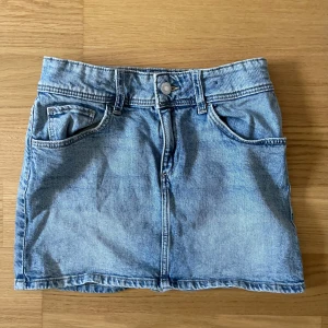 Blå jeanskjol från H&M - Snygg ljusblå jeanskjol från H&M med klassisk femficksdesign, knapp och dragkedja framtill samt hällor för bälte. Kjolen är kort och har en rak passform som ger en cool och avslappnad vibe. Perfekt att matcha med sneakers eller boots.