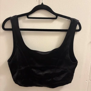 Svart croppad korsettopp i satin från hm - Snygg svart korsettopp med croppad passform och breda axelband. Toppen är sydd i glansigt satinmaterial som ger en lyxig känsla. Perfekt för att lyfta vilken outfit som helst med sin stilrena look och markerade sömmar.