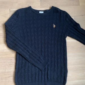 Stickad tröja U.S. Polo  - Säljer en mörkblå stickad tröja från U.S. Polo Assn. med klassiskt kabelstickat mönster och broderad logga på bröstet. Tröjan har rund halsringning och långa ärmar, perfekt för lager-på-lager under kyliga dagar.