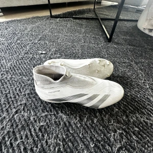 Vita Adidas Predator fotbollsskor - Säljer ett par stilrena vita Adidas Predator fotbollsskor med grå detaljer. Skorna har en strumpliknande passform utan snörning och är tillverkade i syntetmaterial för bästa bollkontroll. Perfekta för dig som vill sticka ut på planen. 