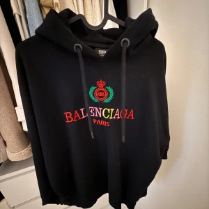 Svart hoodie  - Svart hoodie med broderad logga och emblem i rött, grönt och gult framtill. Tröjan har huva med snörning och en avslappnad passform. Perfekt för dig som vill ha en statement piece med lyxig streetkänsla.